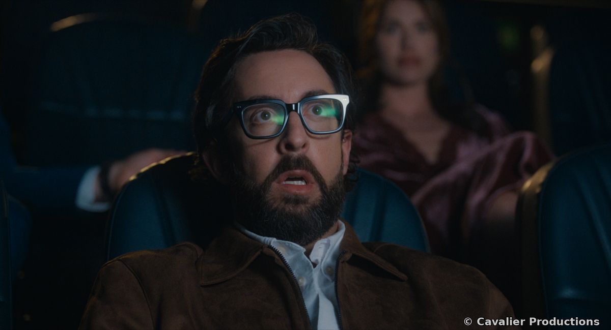 A bearded man in glasses staring, horrified, in a cinema